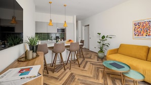 Une cuisine moderne avec des luminaires suspendus, un coin repas doté d’un parquet en chevrons et un canapé jaune dans l’espace de vie.