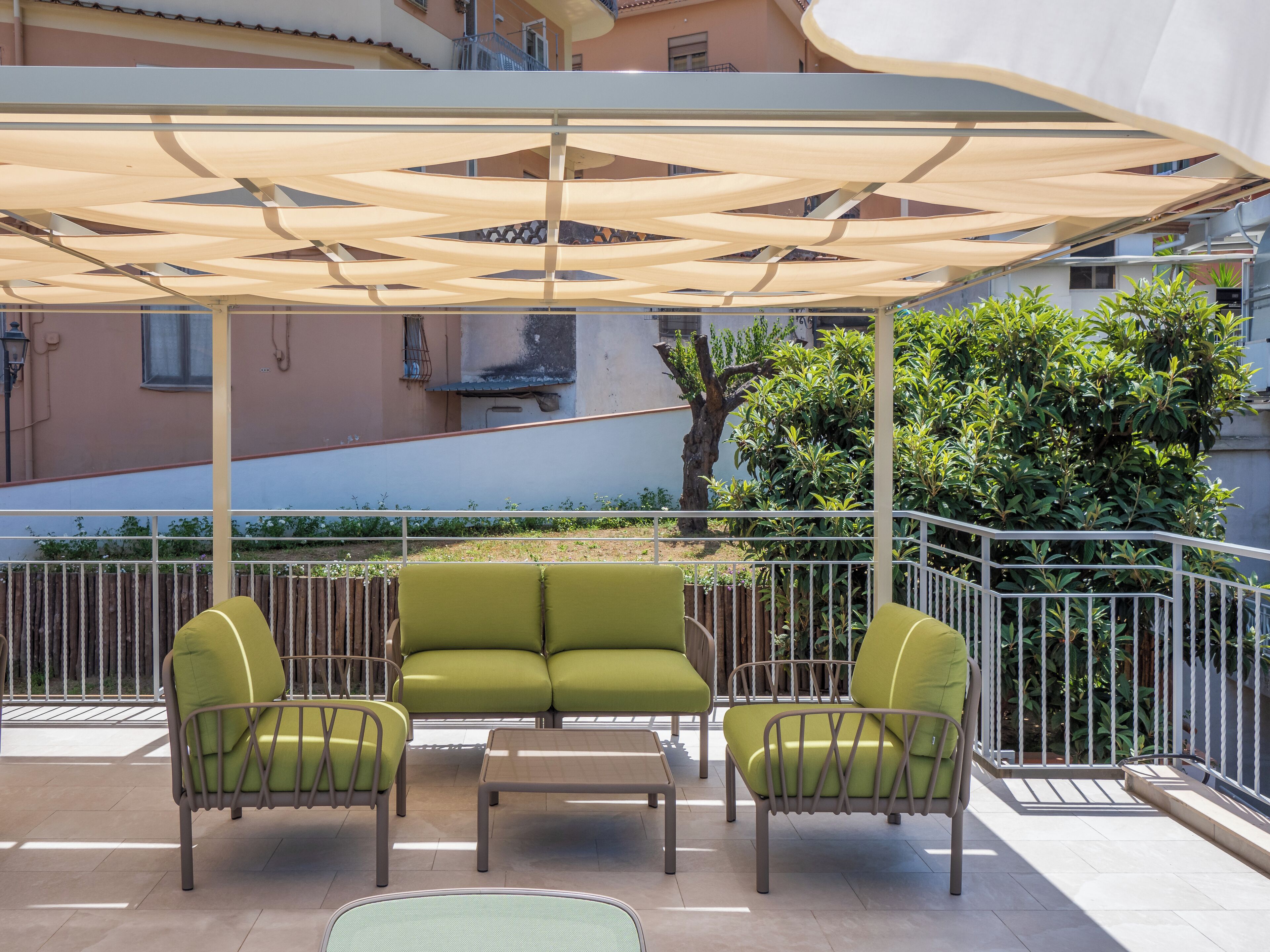An outdoor seating area with a canopy, two green cushioned chairs, a small table, and a railing.