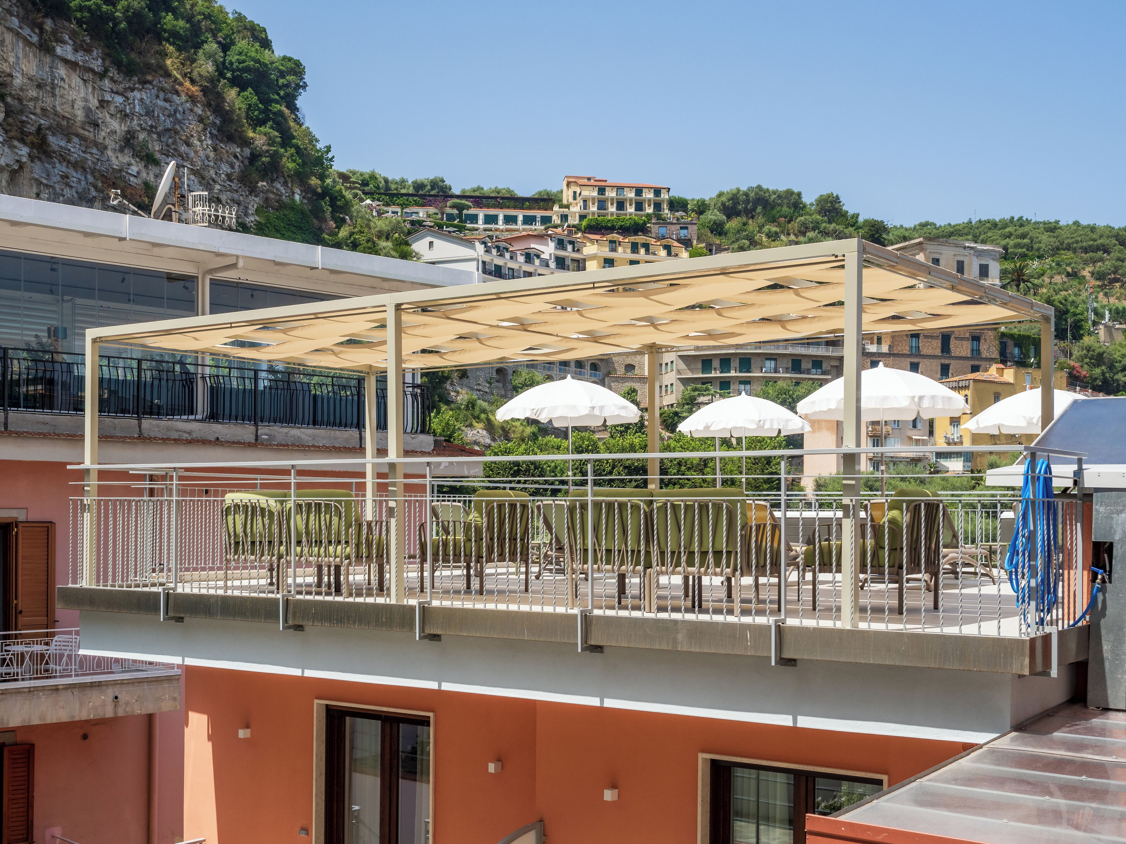 En tagterrasse med udendørs møbler, en pergola og udsigt til bygninger og grønt i baggrunden.