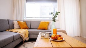 Un salon avec un canapé gris, des coussins jaunes, une table basse sur laquelle se trouve une assiette de pâtisseries, une tasse blanche et un vase avec une plante.