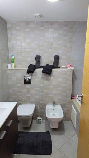 Un baño con inodoro, bidé y lavamanos. Hay toallas en un estante, un dispensador de papel higiénico y un cepillo de limpieza en el suelo.