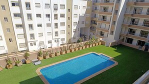 Une piscine entourée d’une pelouse verdoyante, avec un immeuble à plusieurs étages en arrière-plan.