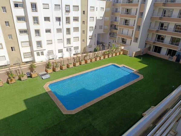 Una piscina rodeada de césped verde, con un edificio de varios pisos al fondo.