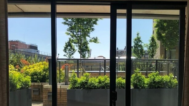 Une pièce dotée d’une porte-fenêtre coulissante donnant sur un patio extérieur, avec un sol carrelé, des jardinières plantées de verdure et une vue sur le paysage urbain, avec ses bâtiments et ses arbres.
