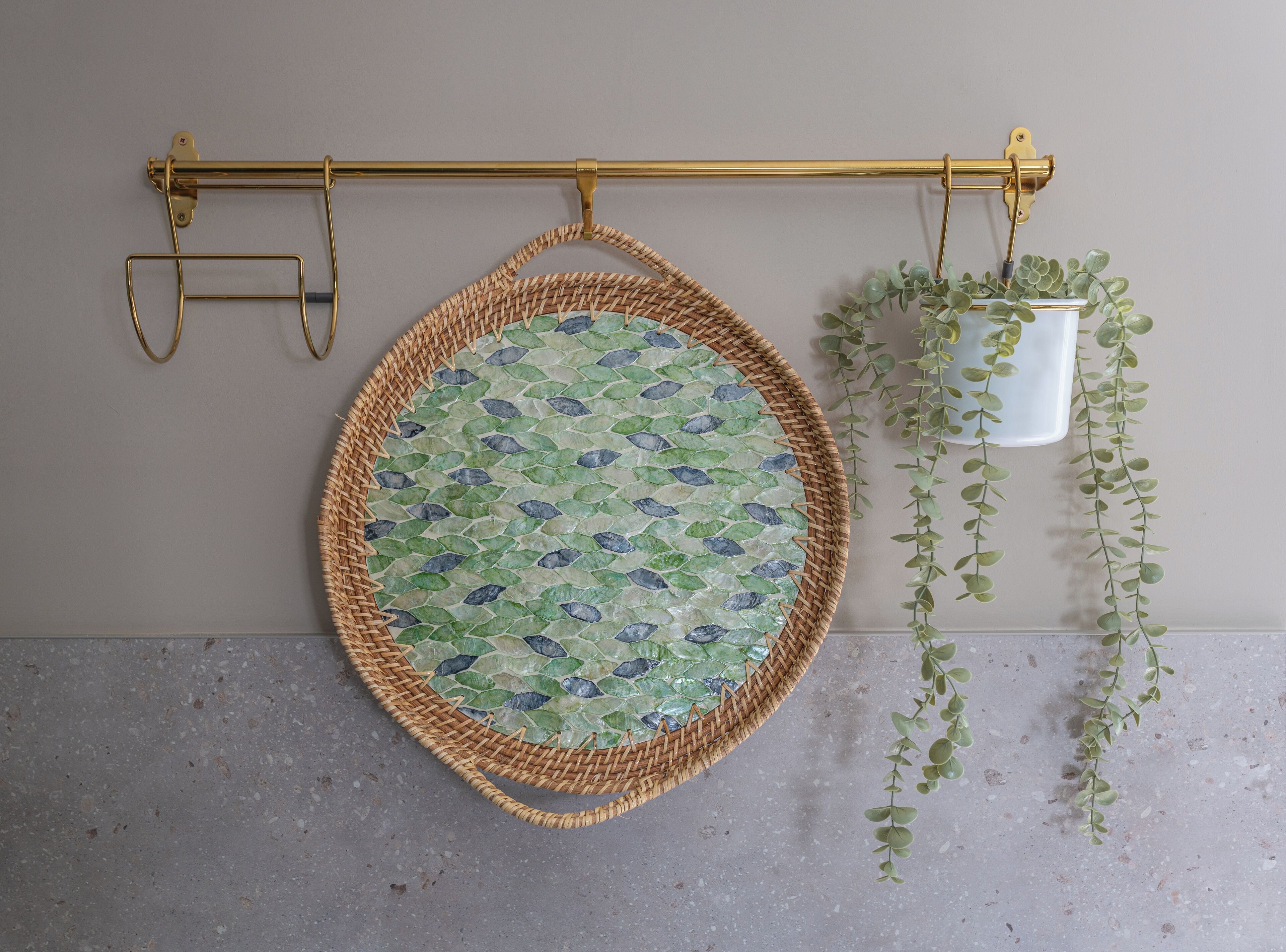 Un panier rond en osier, orné d’un motif en mosaïque, suspendu à une tige dorée, un seau blanc contenant des plantes vertes et un sol en béton.