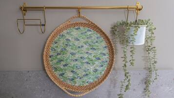 Un panier rond en osier, orné d’un motif en mosaïque, suspendu à une tige dorée, un seau blanc contenant des plantes vertes et un sol en béton.