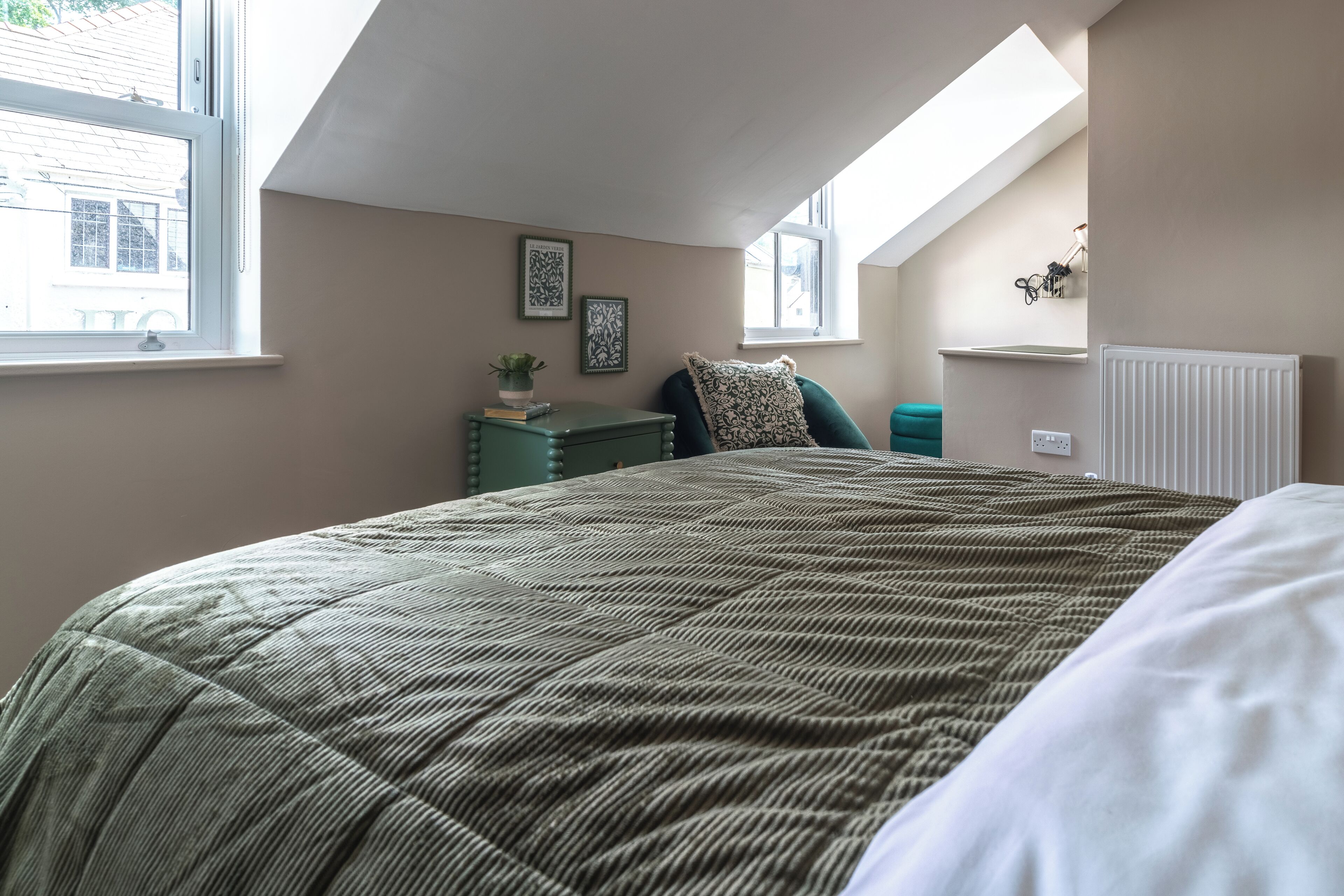 A bedroom with a large bed, a window, a radiator, and a small table with a plant.