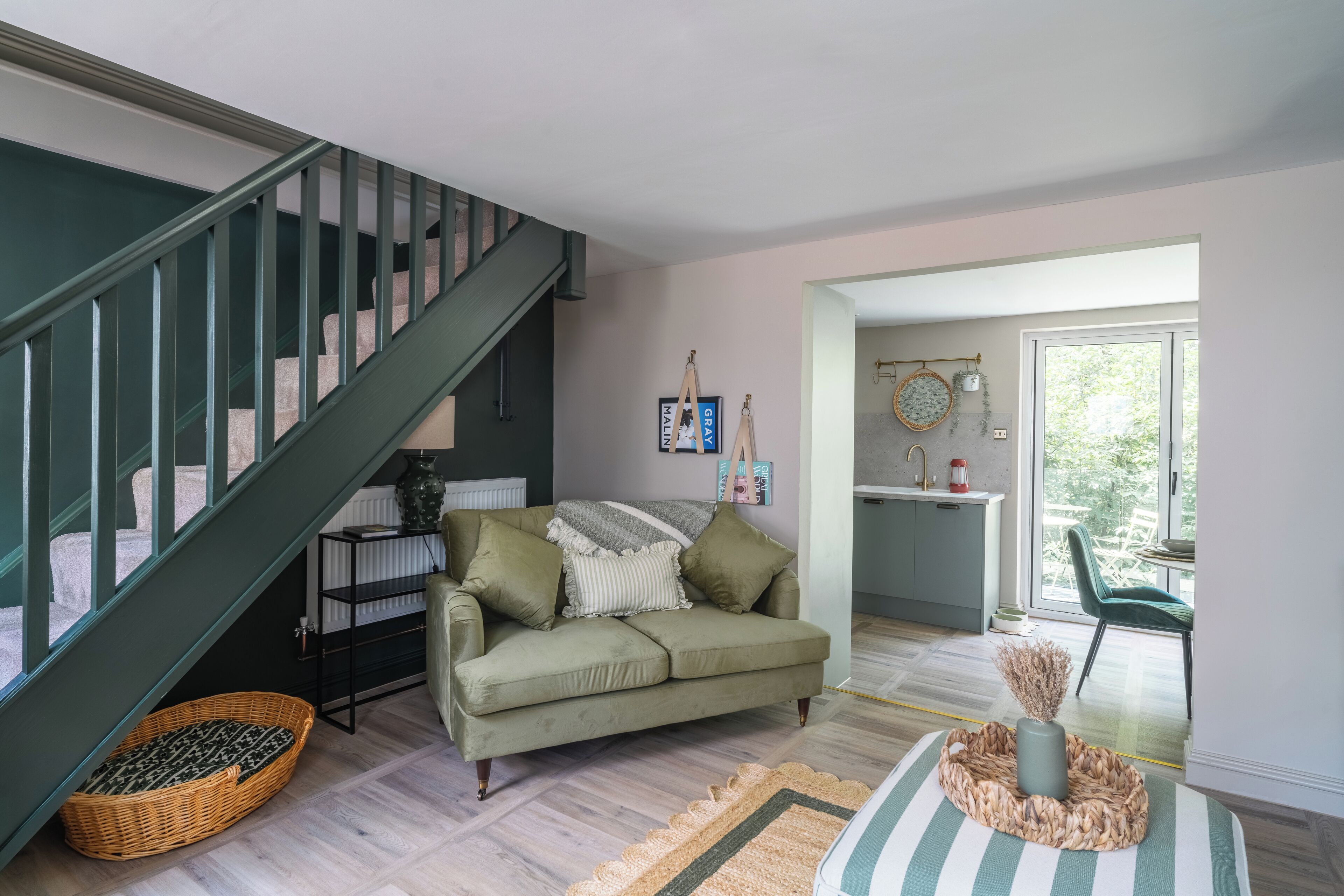 A modern living room with a staircase, a green sofa, a basket, and a kitchen area visible through an open door.
