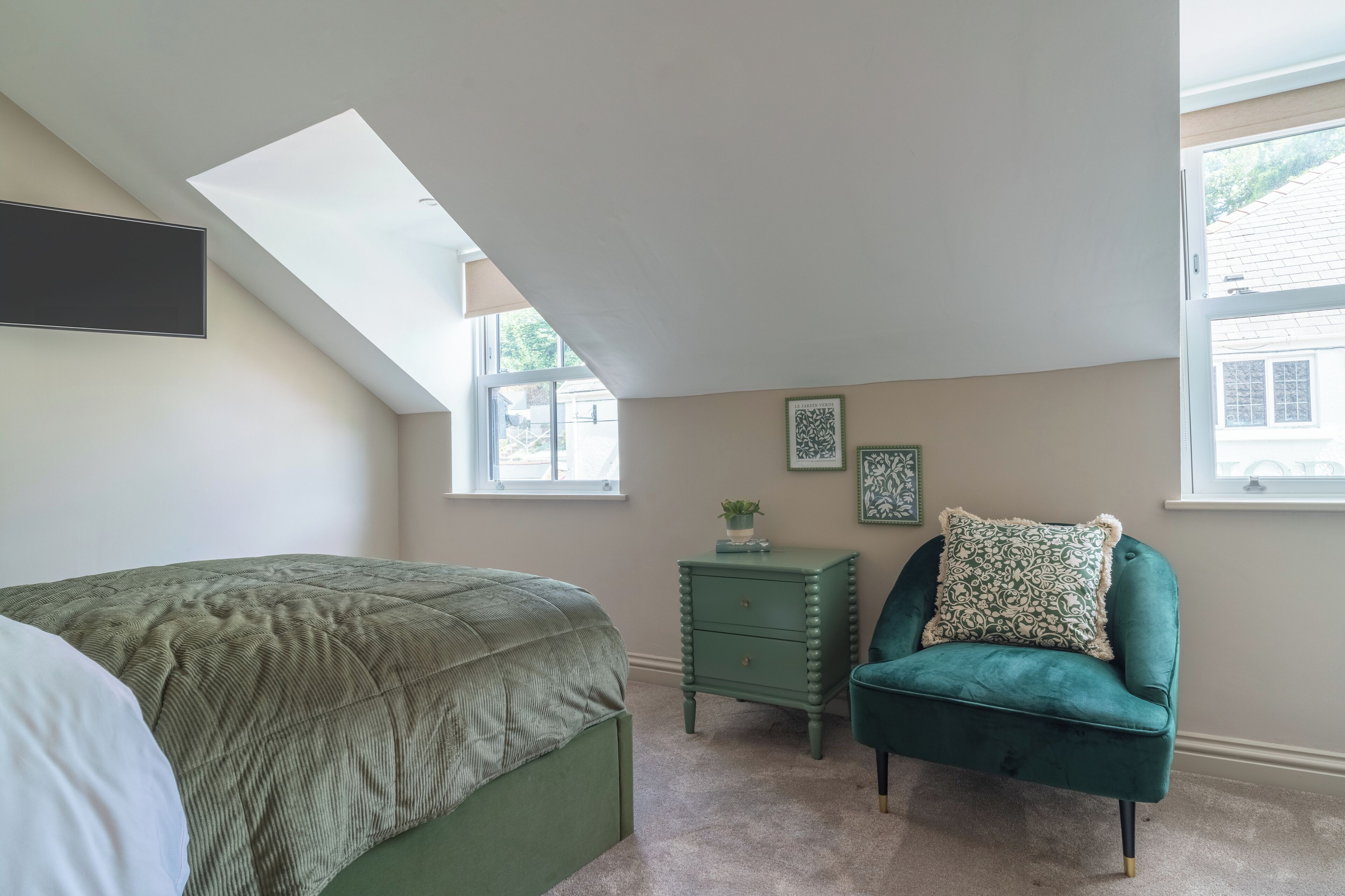 A bedroom with a sloped ceiling, a bed with a green bedspread, a green chair with a patterned pillow, a small green nightstand, and two framed pictures on the wall.