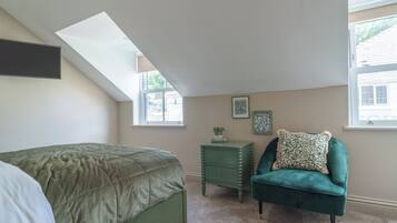 A bedroom with a sloped ceiling, a bed with a green bedspread, a green chair with a patterned pillow, a small green nightstand, and two framed pictures on the wall.