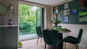 A modern dining area with a glass door leading to a garden, a round table with chairs, and a wall decorated with framed pictures.