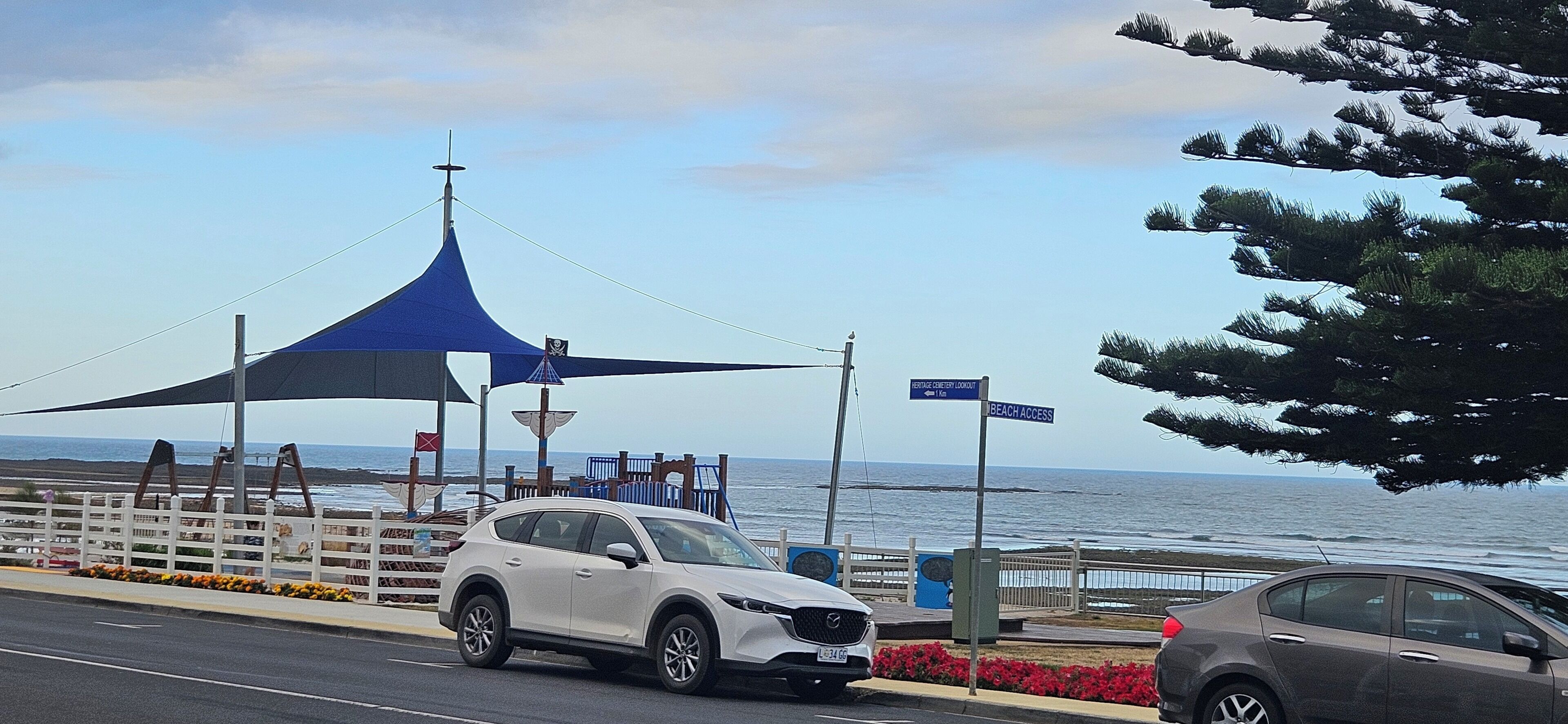 A coastal road with a white SUV parked near a blue canopy structure, a beach access sign, and a view of the ocean.