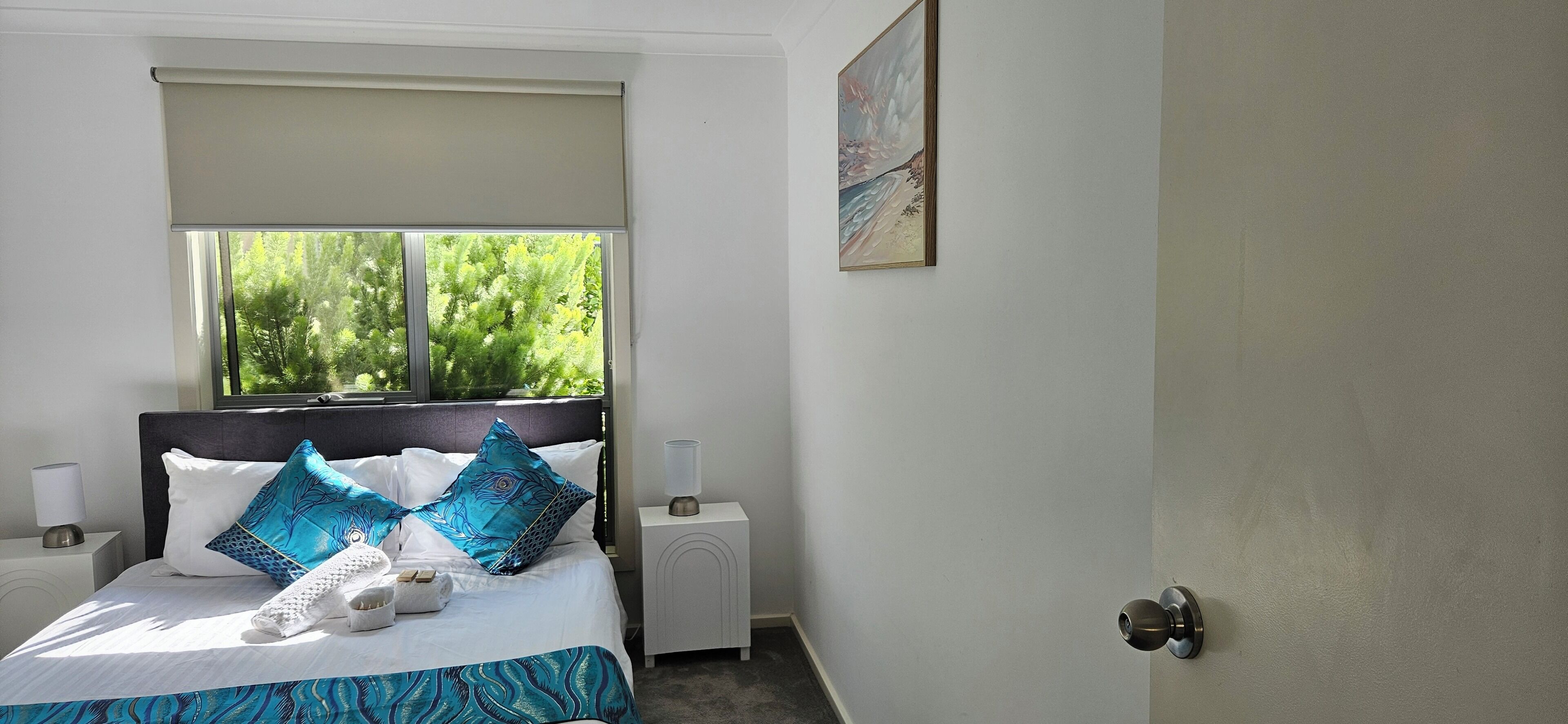 A neatly made bed with a blue and white bedspread, two matching pillows, and a white towel. There is a window with a view of trees and a painting on the wall.
