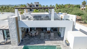 Uma casa moderna de dois andares com um terraço no telhado, piscina e vista para palmeiras e montanhas.