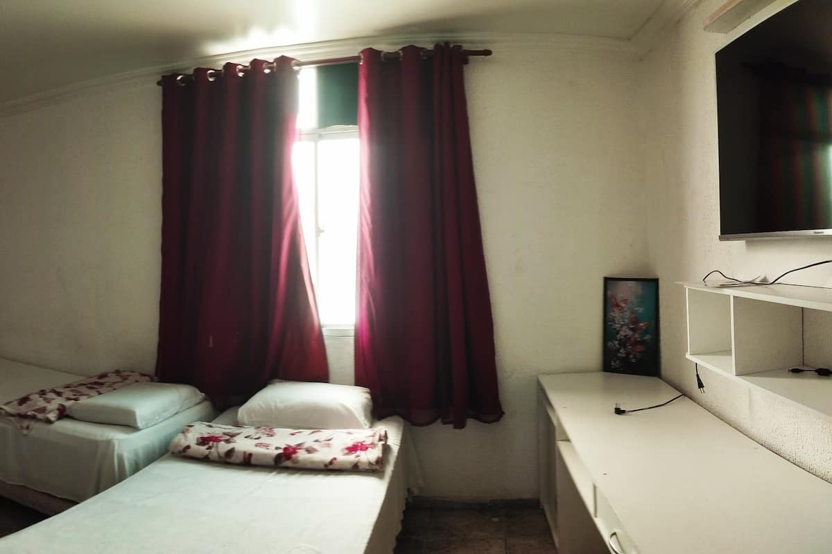 A small, well-lit room with two single beds, a wardrobe, a mirror, and a television.