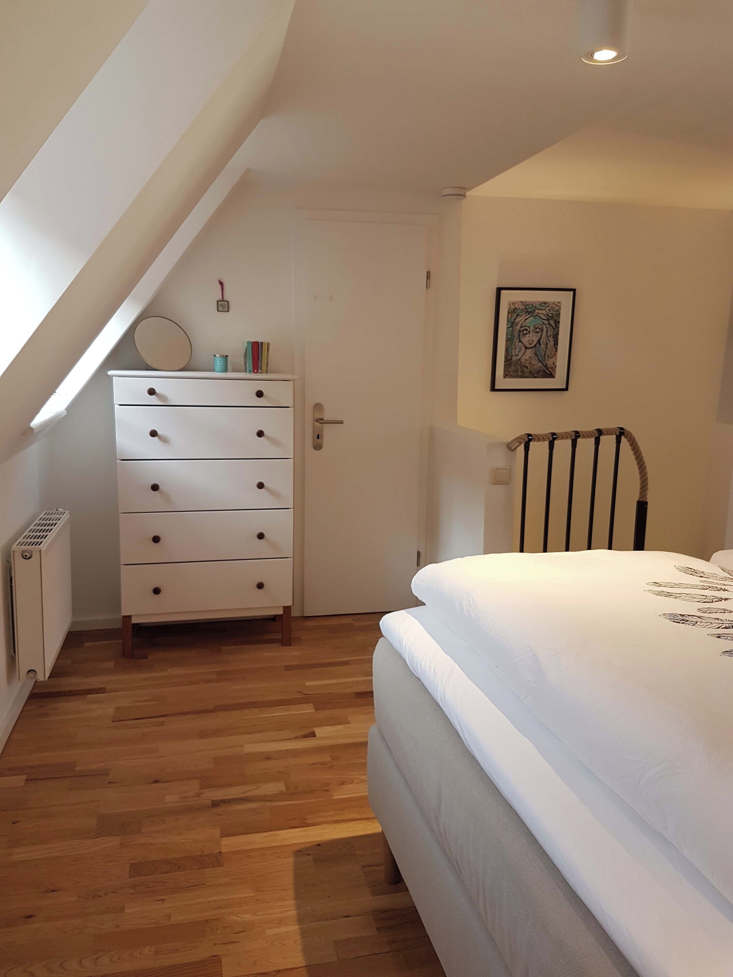 A bedroom with a wooden floor, a bed with a leaf-patterned pillow, a white dresser with drawers, a radiator, a door, and a ceiling light.