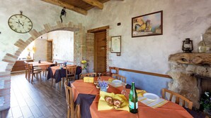 A rustic dining area with brick walls, wooden beams, and a large clock. Tables are set with plates, cutlery, and a bottle of wine.