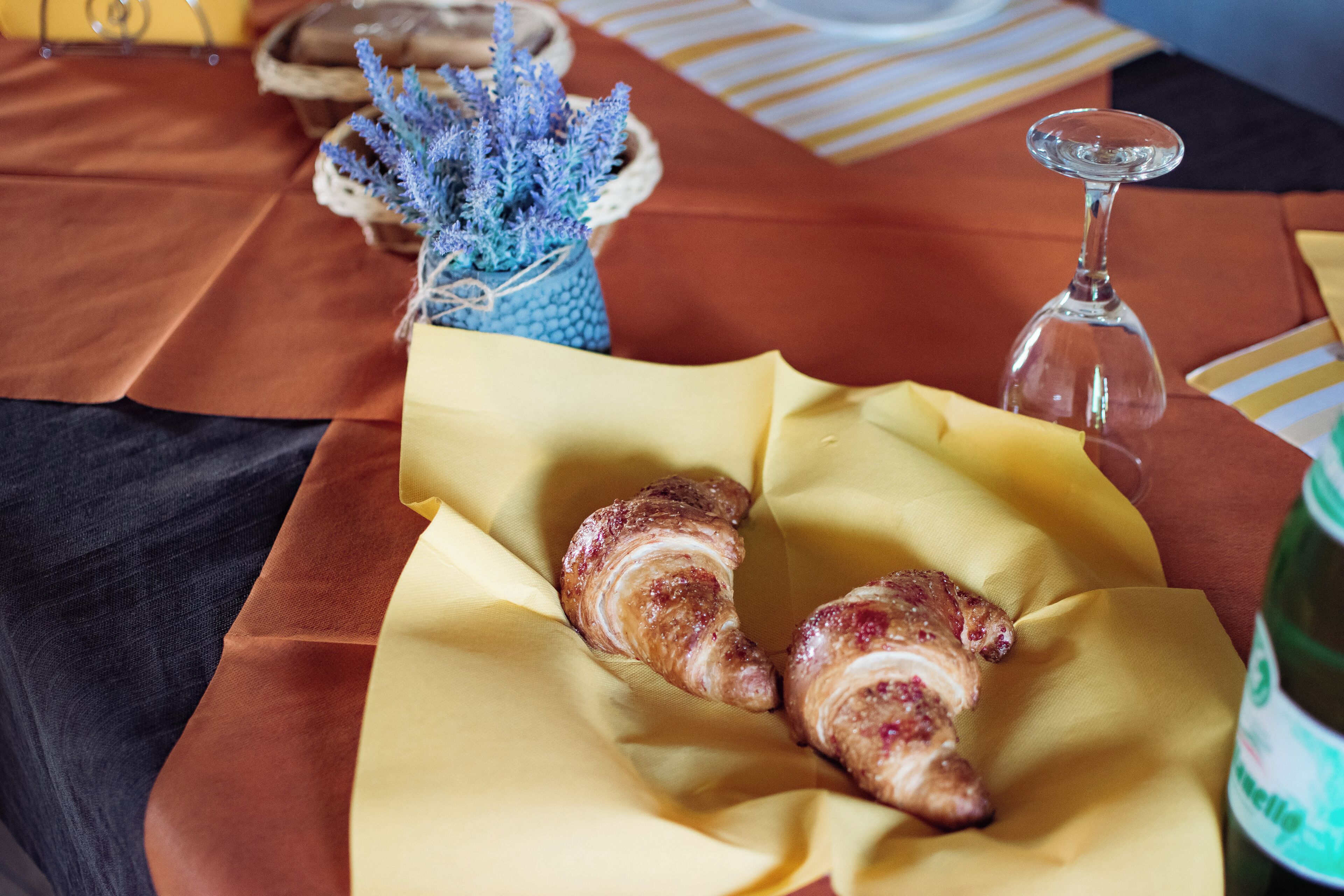 Una mesa con croissants sobre servilletas amarillas, un vaso de agua y un florero azul con lavanda.