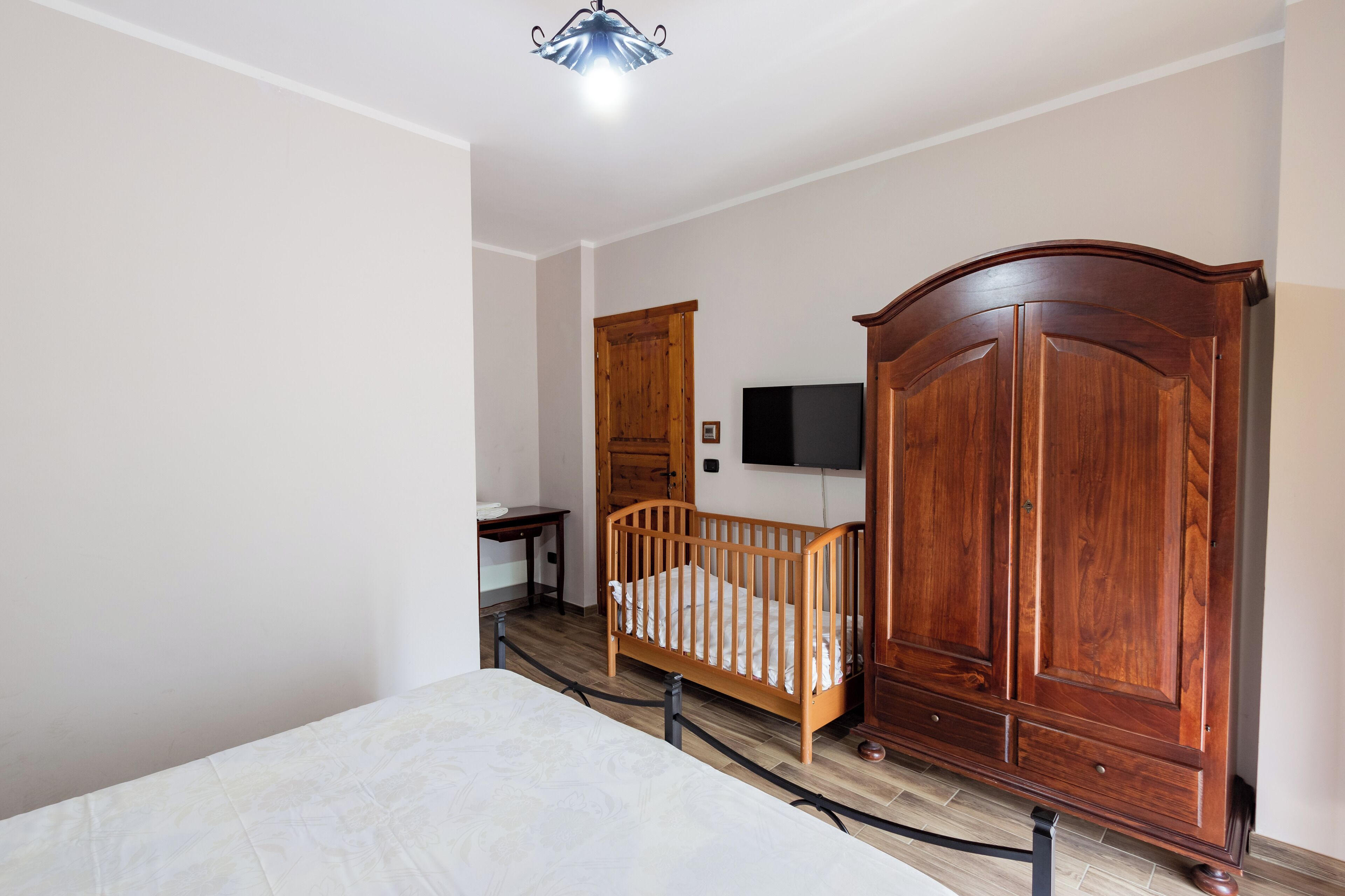 A bedroom with a crib, wooden wardrobe, and a television on the wall.