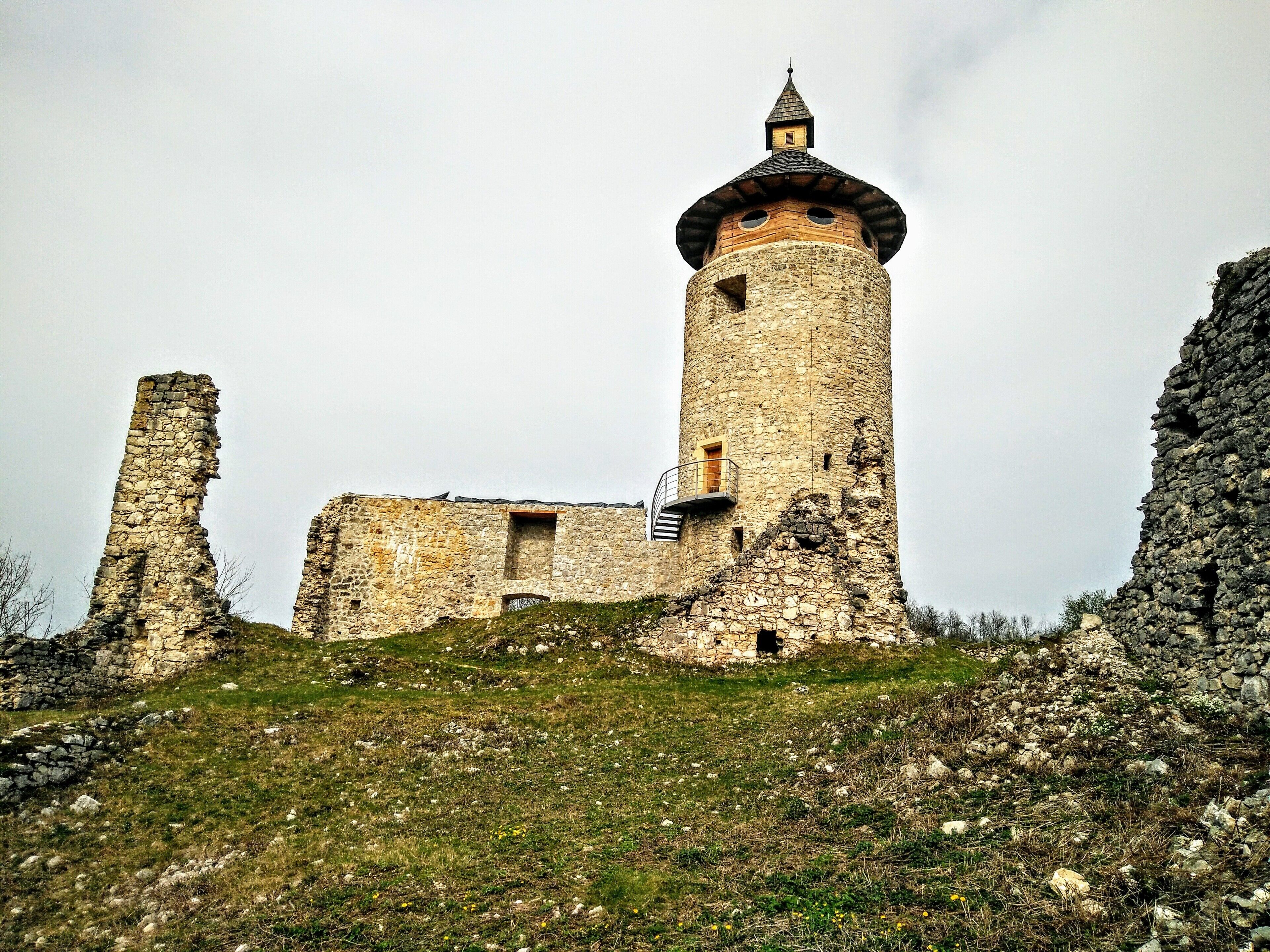 Ein uralter, steinerner Turm mit spitzem Dach, umgeben von Ruinen und grüner Landschaft.