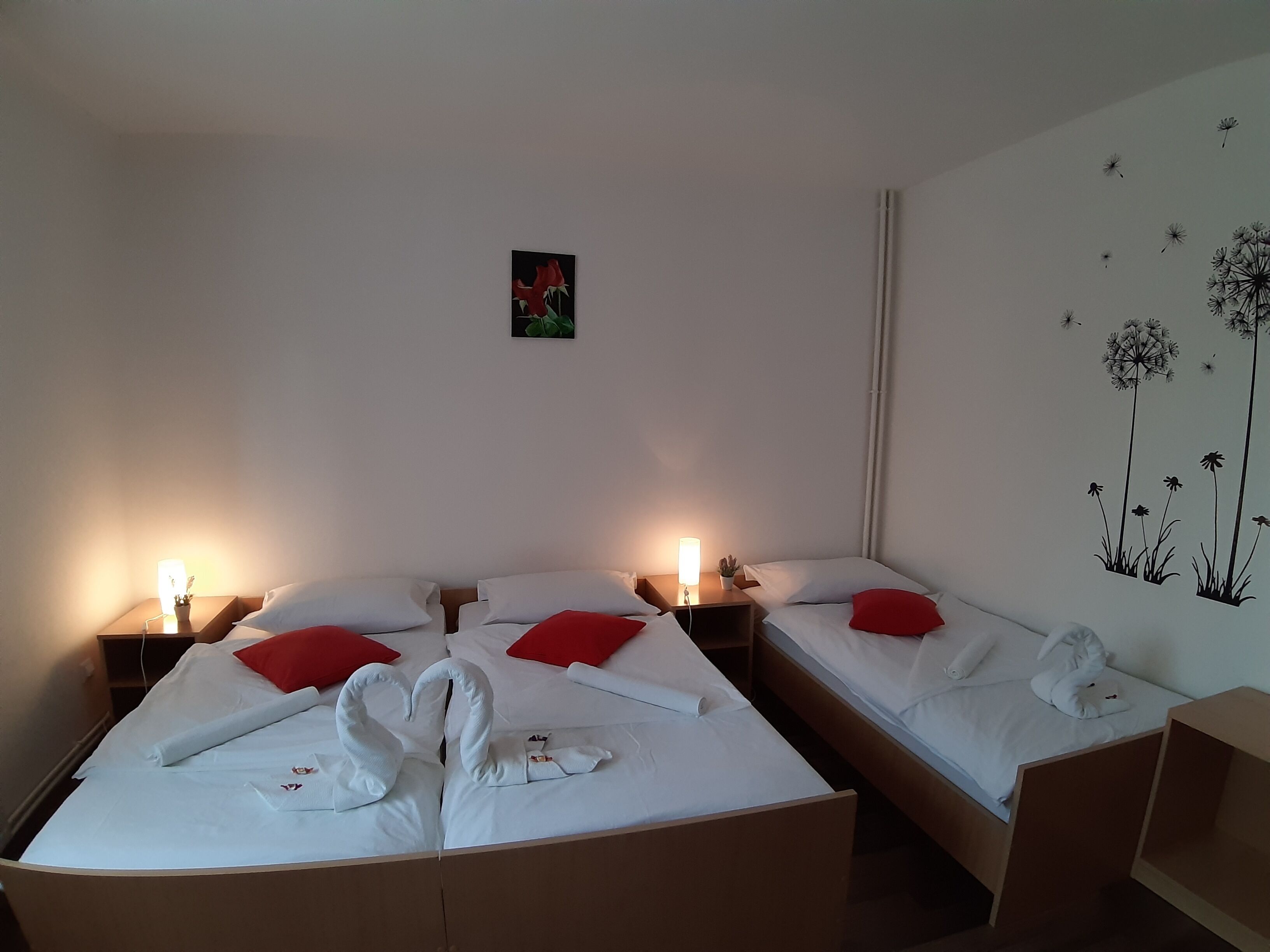 Quarto de hotel com duas camas, cada uma com roupa de cama branca e almofadas vermelhas. Há mesas de cabeceira com abajures, uma pequena planta em uma das mesas, e uma decoração de parede com dentes-de-leão.