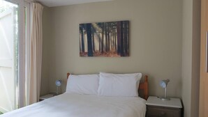 Una habitación de hotel con una cama grande, dos mesitas de noche con lámparas, un cuadro de un bosque en la pared y una ventana con cortinas.