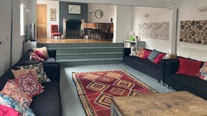 Amplia sala de estar con un sofá grande, una mesa de centro y una alfombra estampada. Se ve una escalera que sube a un piso superior, y un piano al fondo.