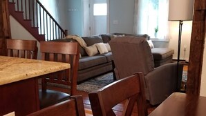 A dining area with wooden chairs and a table, a living room with a sofa and a lamp, and a staircase in the background. - LAKEFRONT COMFORT (Chazy)