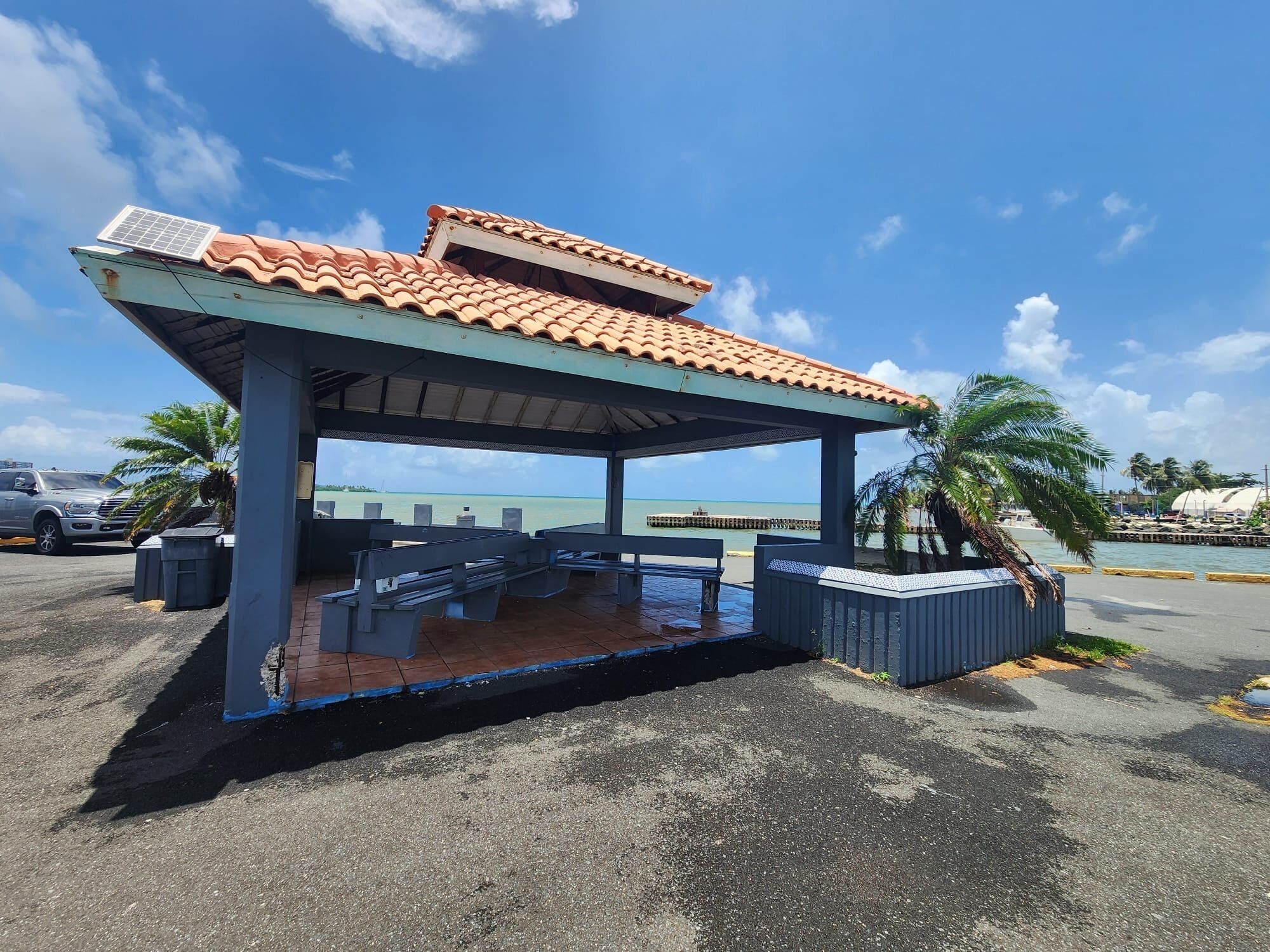Un espacio al aire libre cubierto con bancos y techo de baldosas, con vista a un cuerpo de agua y un muelle.