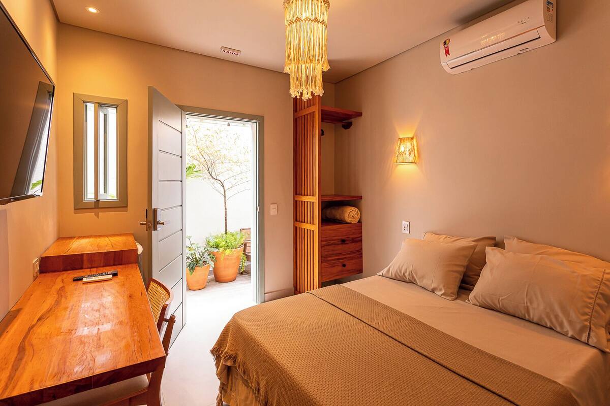Quarto com escrivaninha de madeira, cama com roupa de cama bege, luminária pendente e aparelho de ar condicionado.
