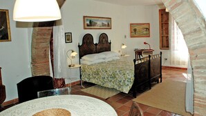 Um quarto com uma cama, duas luminárias de cabeceira, uma mesa de cabeceira, uma cadeira, uma mesa com um jogo americano e um quadro na parede.