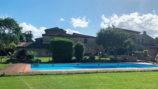 Uma piscina com deck de tijolos, cercada por grama bem cuidada e árvores, com edifícios de pedra ao fundo.