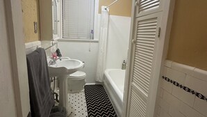 Ein Badezimmer mit einem weißen Waschbecken, einer Toilette und einer Badewanne. Über dem Waschbecken hängt ein Spiegel, und ein Duschvorhang ist teilweise sichtbar. Der Boden ist mit einem schwarz-weißen Teppich bedeckt.