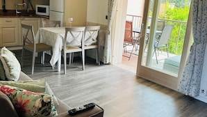 Una sala acogedora con sofá, mesa de comedor y sillas. Se observa una zona de cocina con armarios y electrodomésticos. Un balcón con vistas a la vegetación es visible a través de puertas de vidrio.