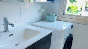 Un baño moderno con un lavabo blanco, tocador de madera oscura, lavadora y ventana con vista a la vegetación.