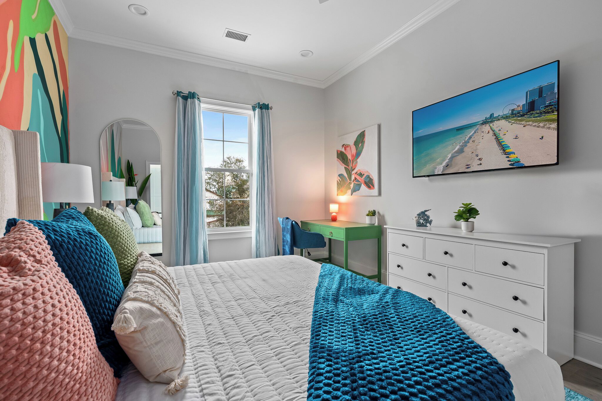 A bedroom with a bed, a desk, a dresser, a mirror, and a painting of a beach.