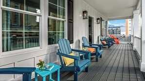 Une véranda couverte avec des fauteuils Adirondack et une table, de grandes fenêtres et une vue sur les bâtiments en arrière-plan.