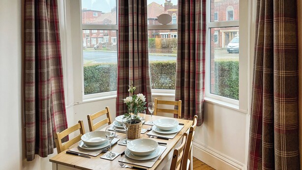 A dining table set for four with a vase of white flowers, wooden chairs, and a view of a residential street through large windows. - Treasure Cove-UK48780 (Whitby)