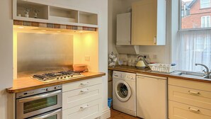 A compact kitchen with a gas stove, oven, washing machine, and a window with a view of a house. - Treasure Cove-UK48780 (Whitby)