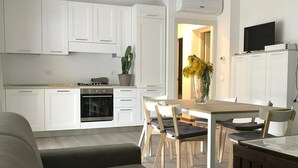 Een moderne keuken met witte kasten, een eethoek met een houten tafel en stoelen, en een grijze bank op de voorgrond.