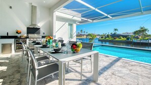 A modern kitchen with a large island, a built-in grill, and a view of a swimming pool. - VRCC Villa Alany- Luxury Getaway- lastminute deals (Cape Coral)