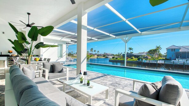 A modern outdoor lounge area with a pool, white furniture, and a view of a residential area. - VRCC Villa Alany- Luxury Getaway- lastminute deals (Cape Coral)