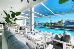 A modern outdoor lounge area with a pool, white furniture, and a view of a residential area.