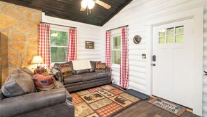 A cozy cabin interior with a stone wall, a ceiling fan, a fireplace, and a checkered rug.