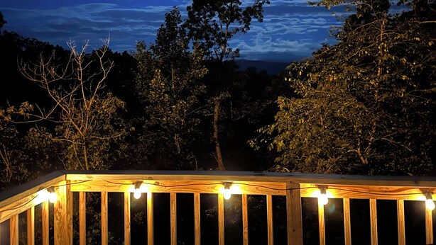 A wooden deck with chairs and string lights, overlooking a forested area with a night sky and visible moon.