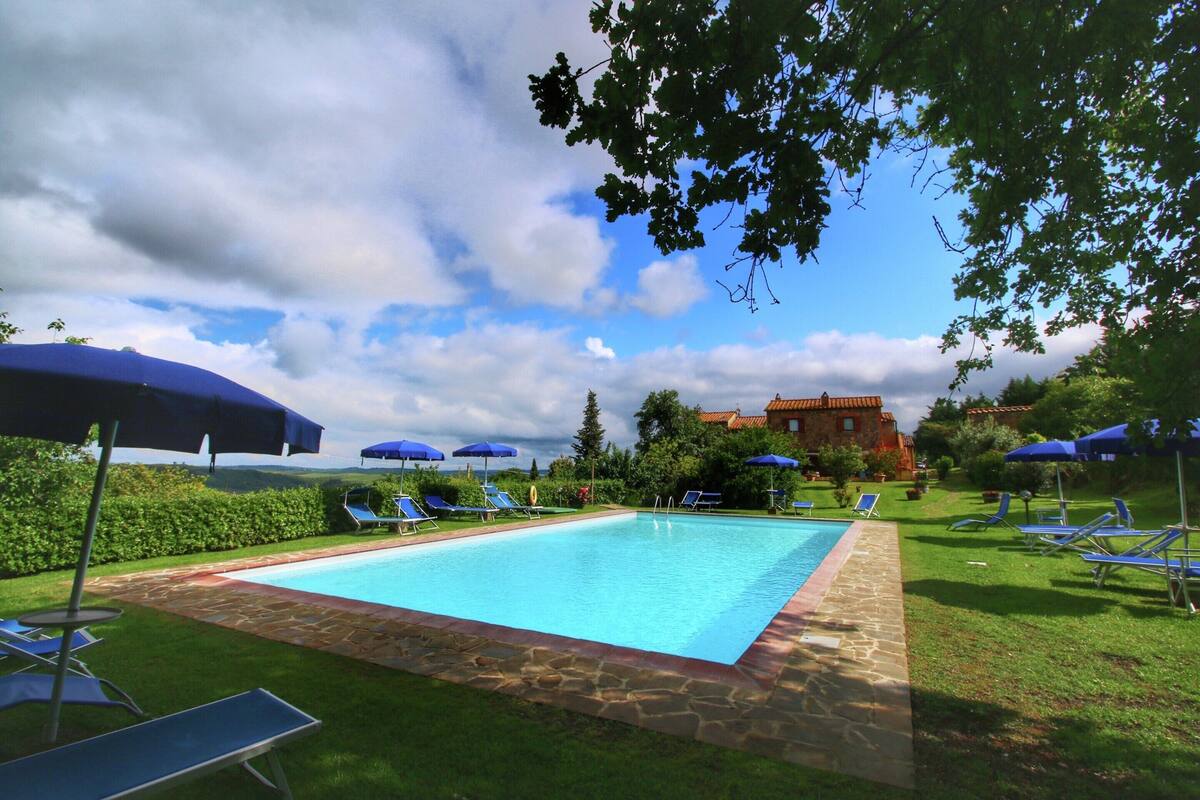 Ein Swimmingpool mit blauen Liegestühlen und Sonnenschirmen, umgeben von einer Steinmauer und viel Grün.