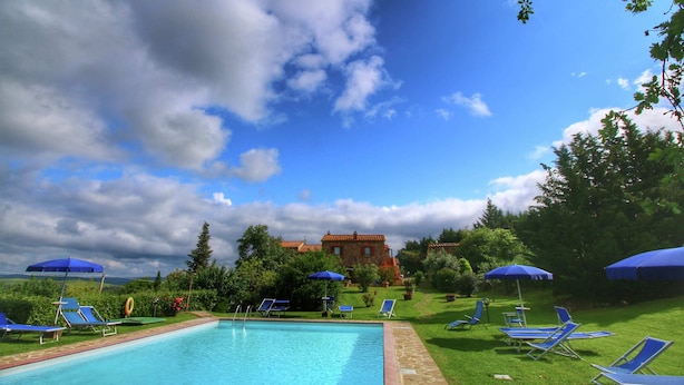 A swimming pool with lounge chairs and umbrellas, surrounded by greenery and a building in the background.