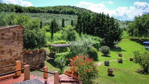 A stone building with a wooden staircase, a well-maintained garden with various plants and a red flowering bush, and a clear view of a forested hill in the background.