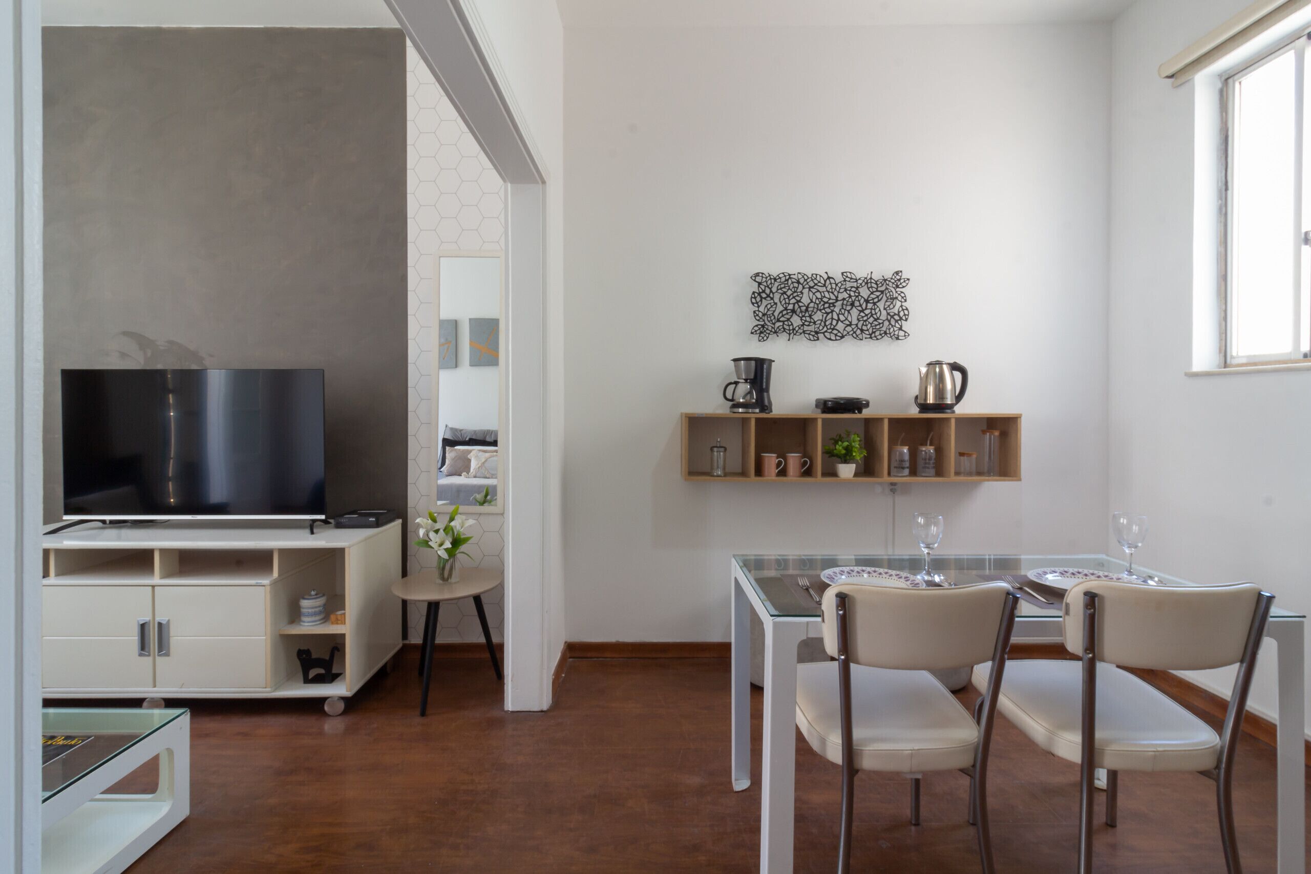 A modern kitchen with a built-in TV, a dining area with a glass table and chairs, and a shelf with kitchenware.