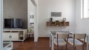 A modern kitchen with a built-in TV, a dining area with a glass table and chairs, and a shelf with kitchenware. - Refuge in Copacabana for two | NSC1011 (Rio de Janeiro)